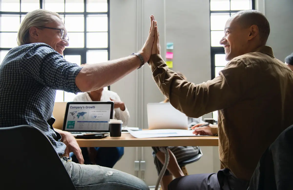 photo of two coworkers sitting at desk and high-fiving because they reached their goals. 